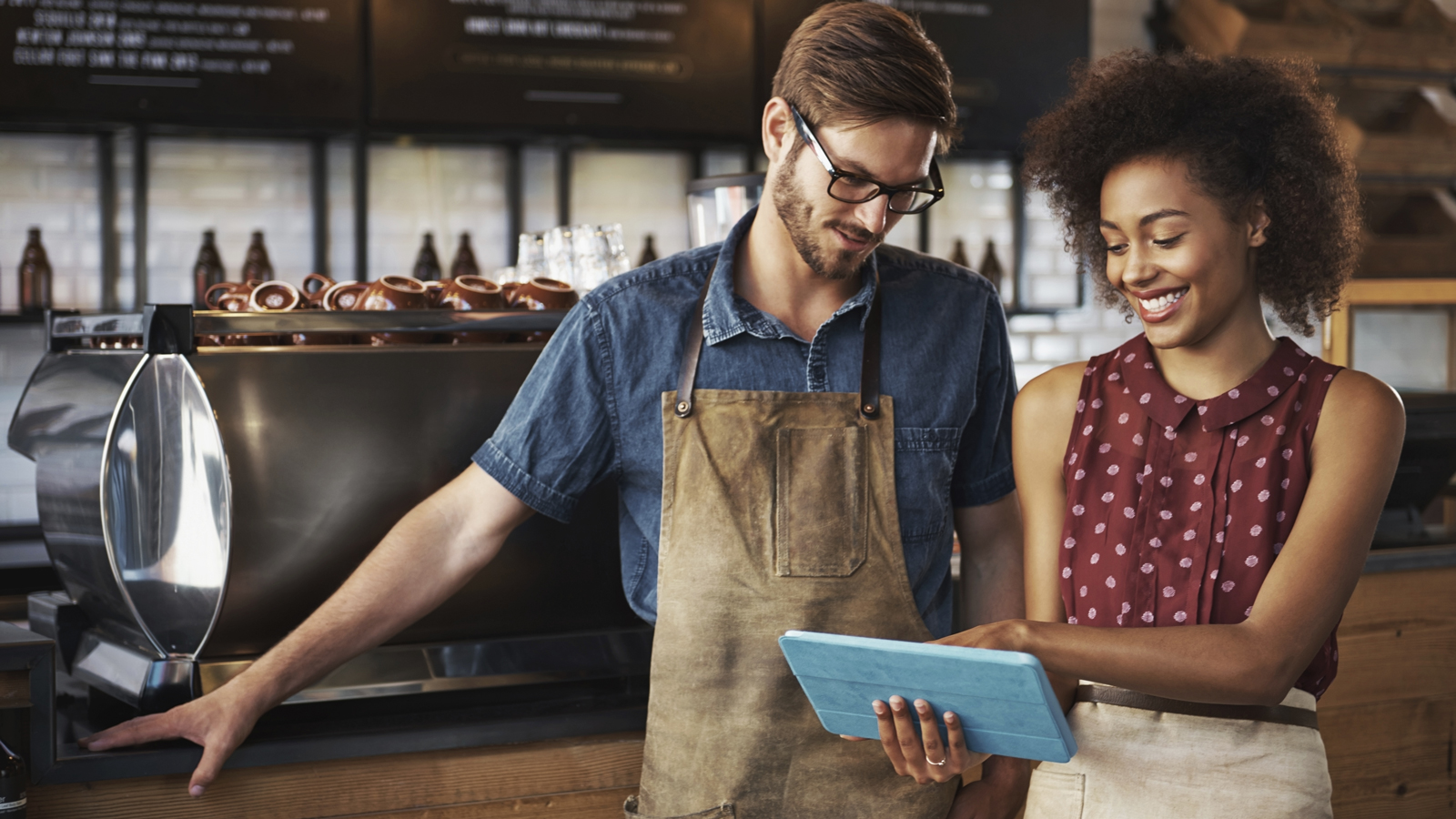 A smiling woman and man standing inside a coffee shop looking at a digital tablet.