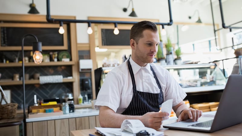 Man in an apron working on a laptop at the front counter of his shop.