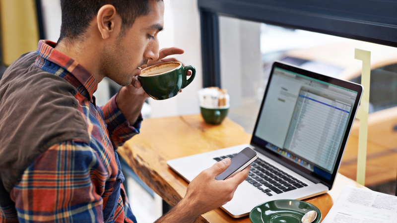 Man at a coffee shop sipping a cup of coffee in front of his laptop.